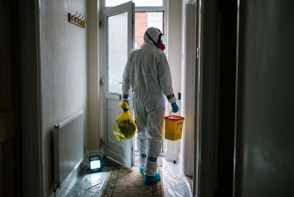 A specialist in protective equipment carrying a sealed clinical waste bag and a sharps container down a hallway towards the front door